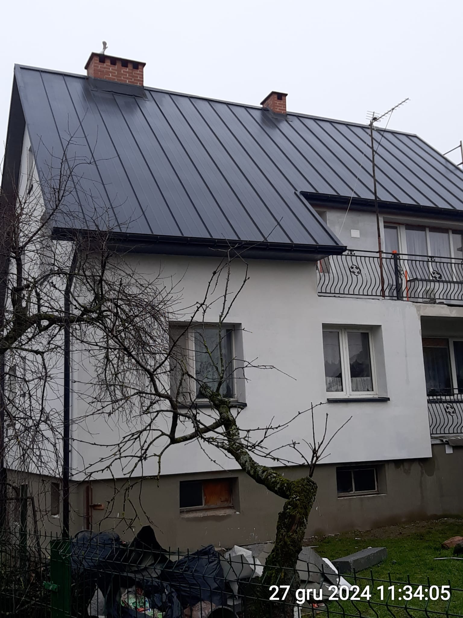 Dwukondygnacyjny dom z nowym, ciemnym dachem z blachy trapezowej i balkonem z ozdobną balustradą, widziany zza drzewa bez liści.