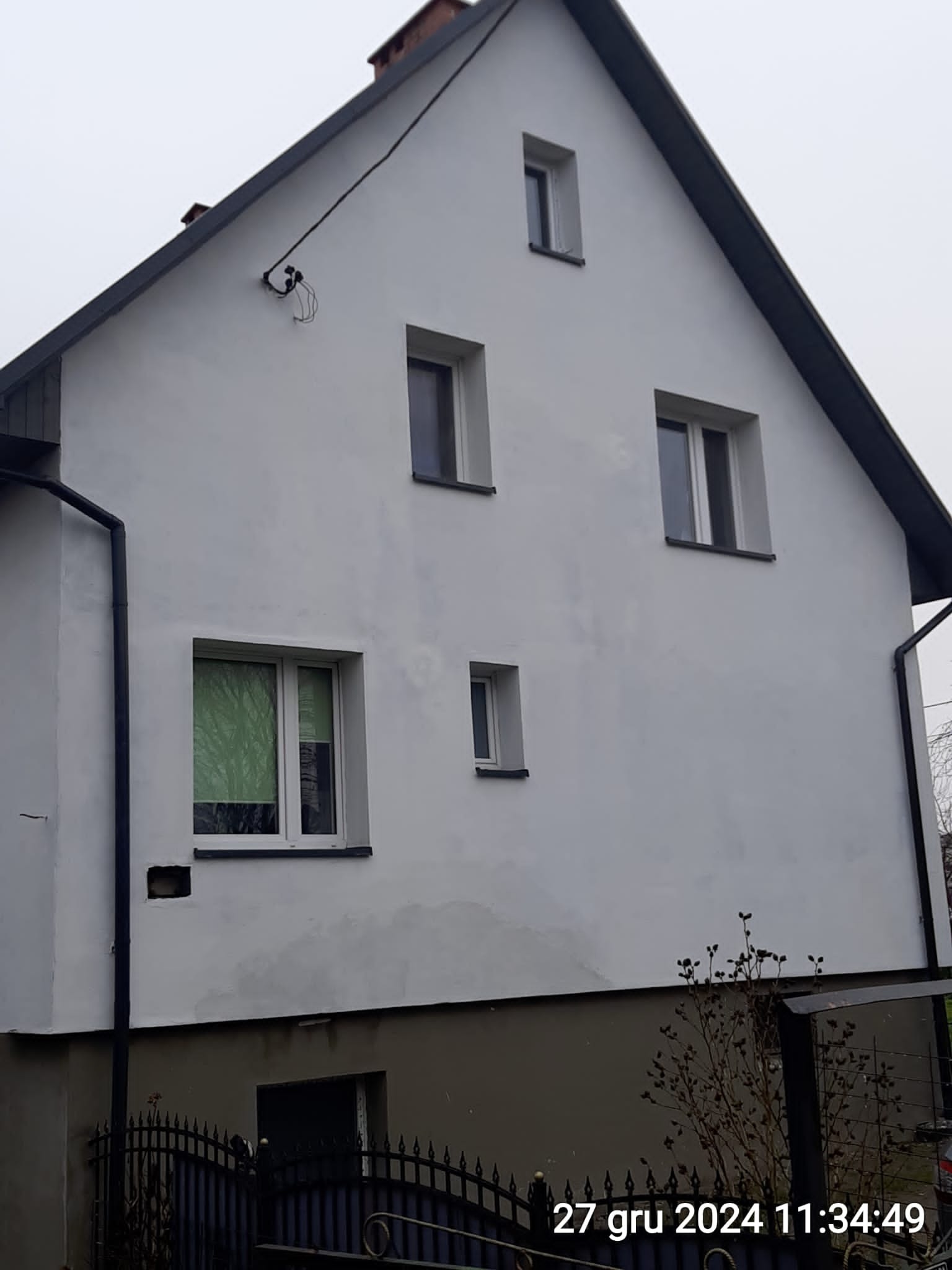 Biała fasada dwukondygnacyjnego domu z kilkoma oknami o różnych rozmiarach, widoczne rynny i instalacja elektryczna na ścianie. Ujęcie z dołu, pochmurne niebo.