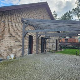 BARWOOD CIESIELSTWO PAWEŁ BARANOWSKI - Murowany dom z cegły klinkierowej z zadaszeniem tarasowym z ciemnego drewna, widoczne rusztowanie, kostka brukowa na podjeździe i trawnik z tyłu.