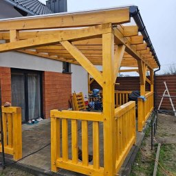 BARWOOD CIESIELSTWO PAWEŁ BARANOWSKI - Świeżo pomalowana na żółto drewniana pergola z barierkami na wybrukowanym tarasie, widok na dom z ceglaną elewacją i sprzęt budowlany w tle.