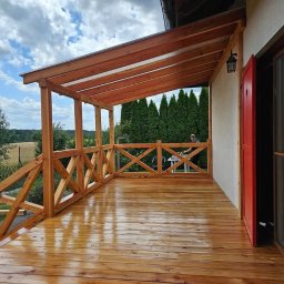 BARWOOD CIESIELSTWO PAWEŁ BARANOWSKI - Zadaszony taras z jasnego drewna z balustradą w kształcie krzyży, świeżo polakierowana podłoga odbija światło, w tle widoczny mężczyzna przy stoliku oraz rząd wysokich tuj.