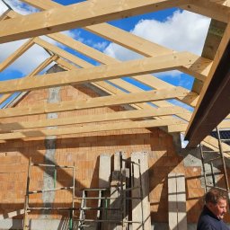 BARWOOD CIESIELSTWO PAWEŁ BARANOWSKI - Wznoszenie drewnianej konstrukcji dachu z widoczną więźbą i elementami murowanej ściany z pustaków ceramicznych, na tle błękitnego nieba z chmurami, z widocznymi rusztowaniami i fragmentem sylwetki...