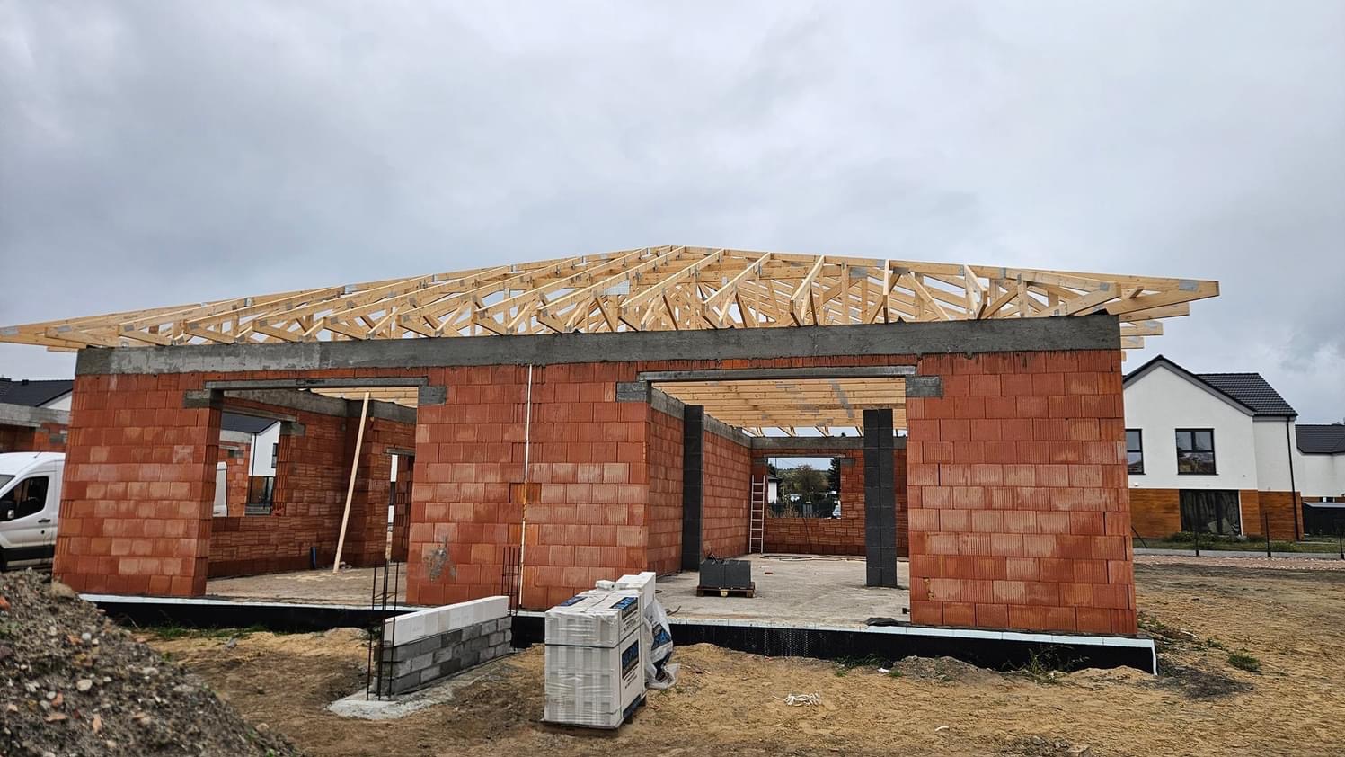 Budowa domu jednorodzinnego w stanie surowym otwartym: czerwone bloczki, betonowy wieniec i drewniana konstrukcja dachu w trakcie montażu, widoczne pustaki i drabina wewnątrz.