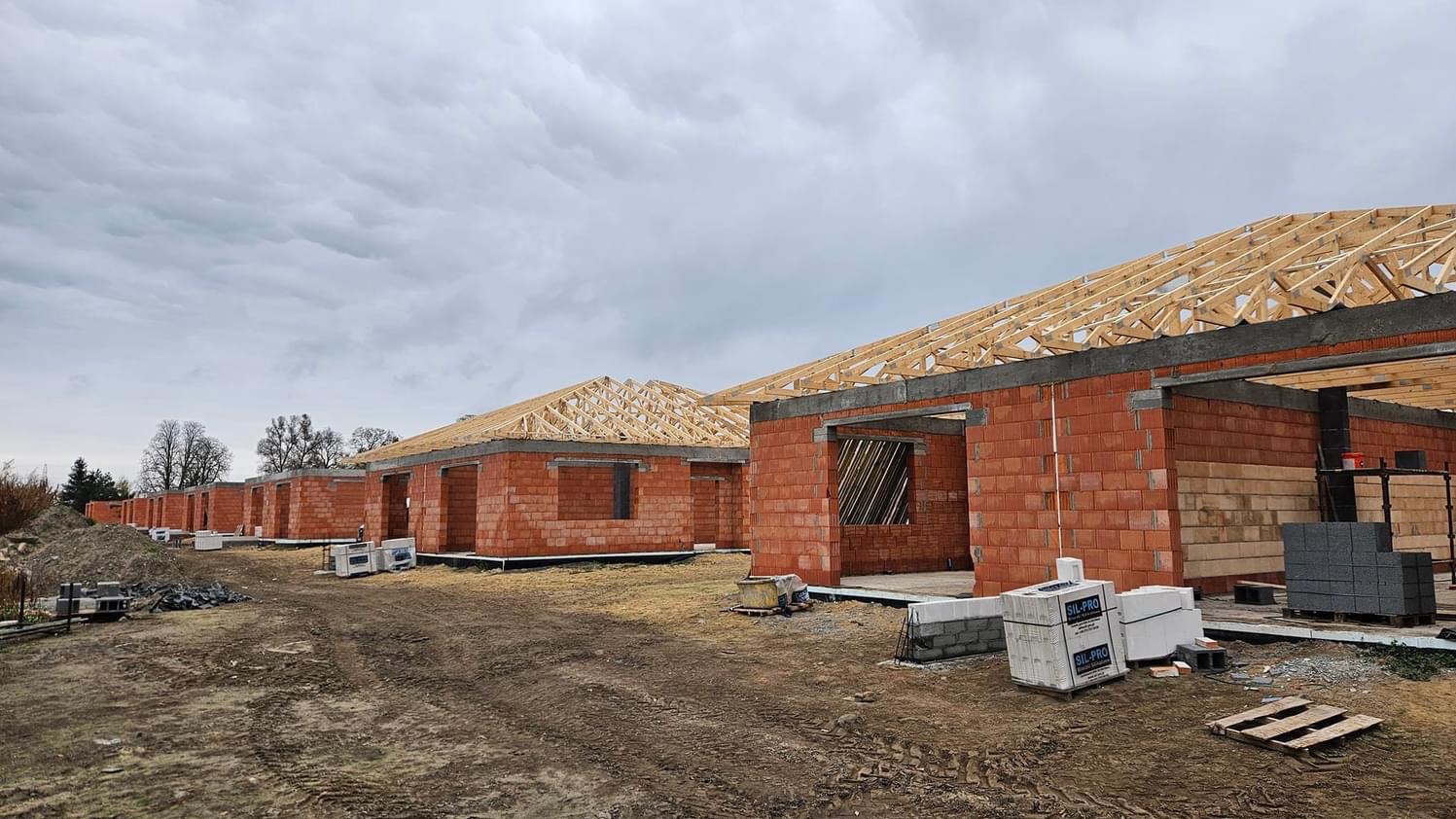 Szereg surowych domów jednorodzinnych w trakcie budowy, z widoczną konstrukcją drewnianą dachu i ścianami z czerwonej cegły, na nierównym terenie z materiałami budowlanymi i pochmurnym niebem w tle.