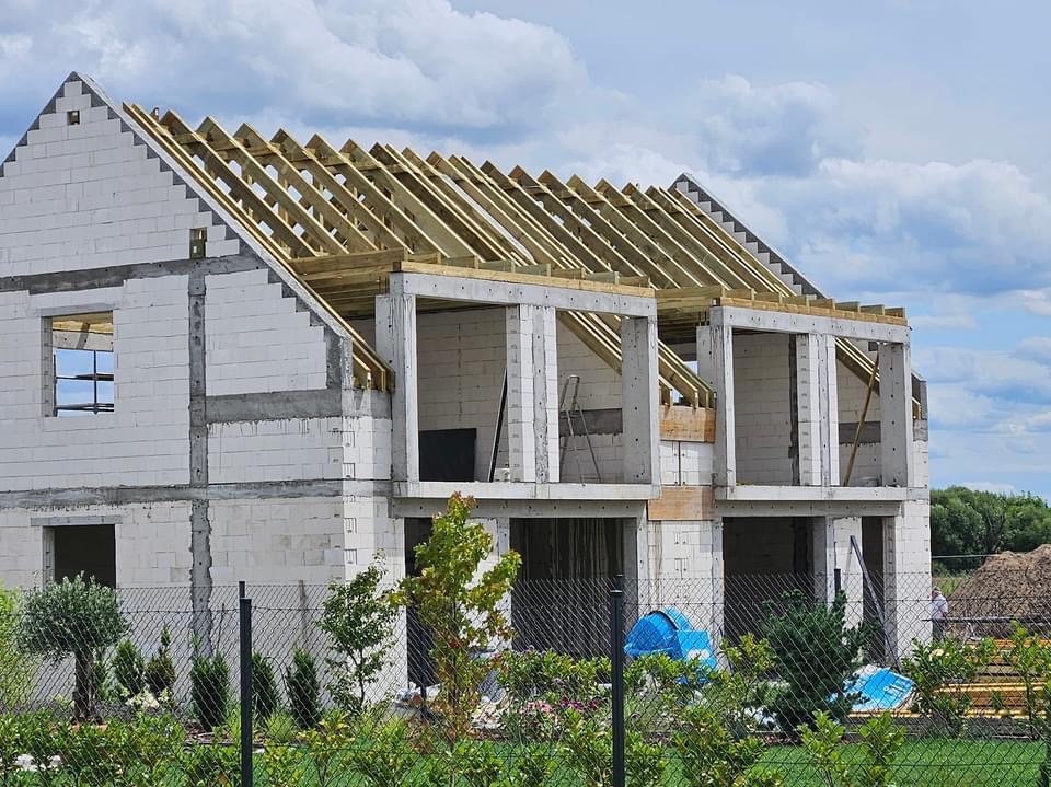 Budowa domu w Murowanej Goślinie: widok na wznoszoną konstrukcję z betonu komórkowego, z widoczną więźbą dachową, zabezpieczoną folią budowlaną i drabina oparta o ścianę.