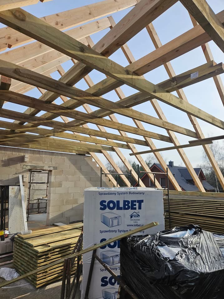 Konstrukcja drewniana dachu w trakcie budowy, widoczne krokwie i murłaty na tle nieba, w tle budynek w stanie surowym i materiały budowlane Solbet.
