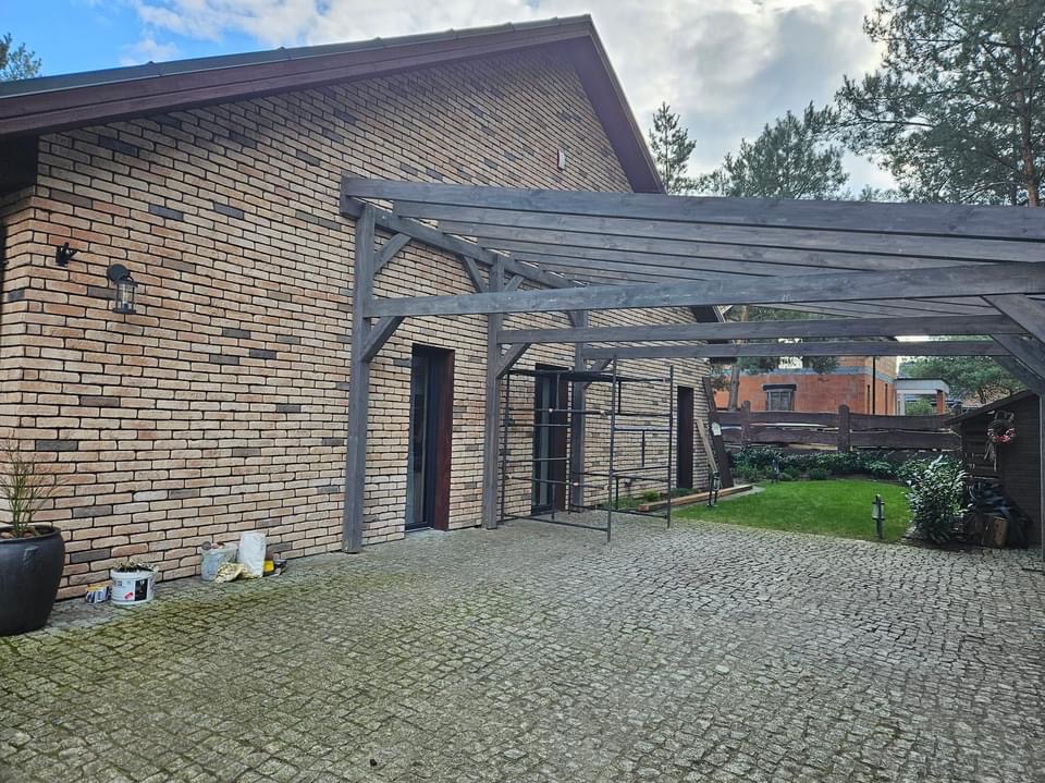 Murowany dom z cegły klinkierowej z zadaszeniem tarasowym z ciemnego drewna, widoczne rusztowanie, kostka brukowa na podjeździe i trawnik z tyłu.