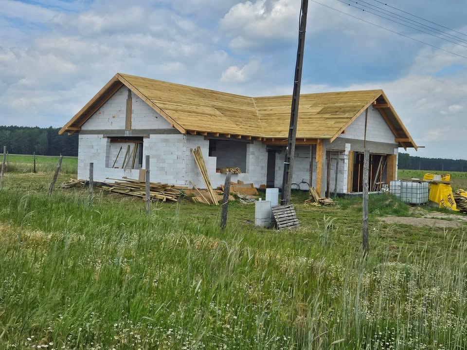 Surowy stan domu jednorodzinnego z bloczków, z częściowo ukończoną więźbą dachową z jasnego drewna, widoczne materiały budowlane i wysoka trawa na pierwszym planie.