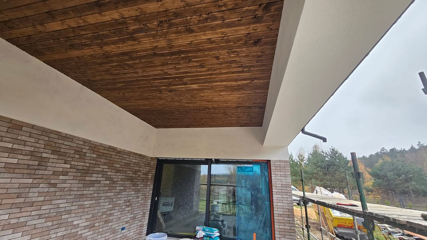 Drewniana podbitka dachowa z widoczną konstrukcją dachu i elewacją z cegły klinkierowej, okno zabezpieczone folią ochronną, rusztowanie w tle.