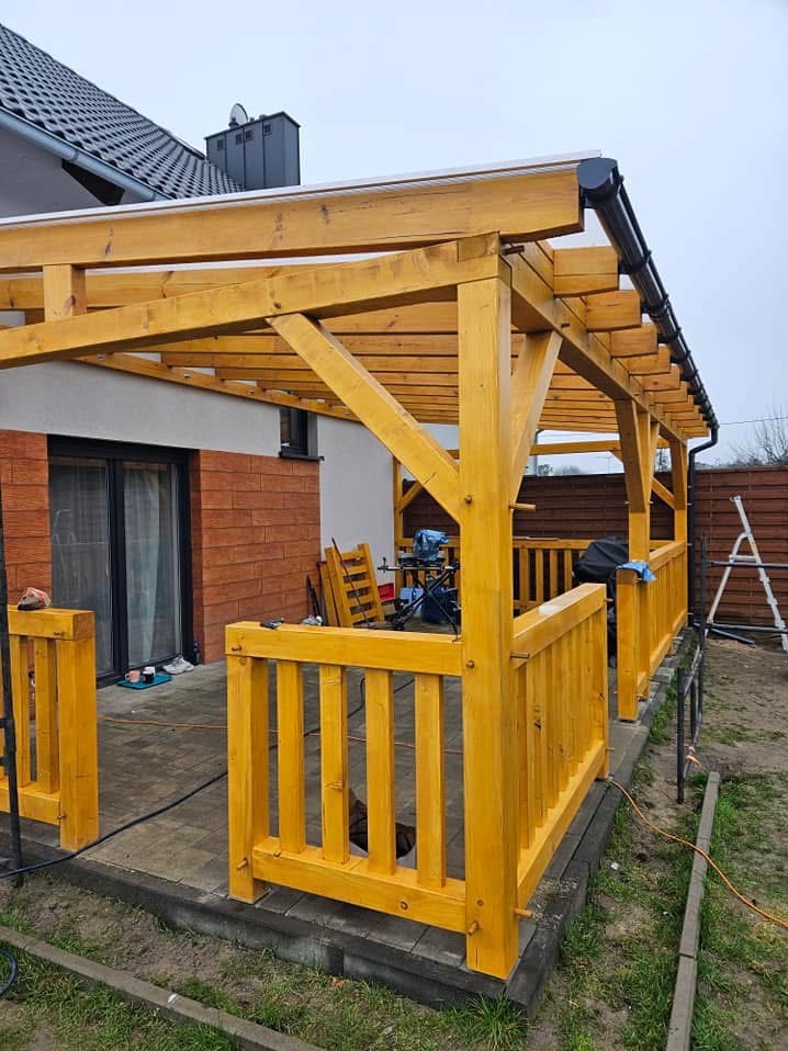 Świeżo pomalowana na żółto drewniana pergola z barierkami na wybrukowanym tarasie, widok na dom z ceglaną elewacją i sprzęt budowlany w tle.