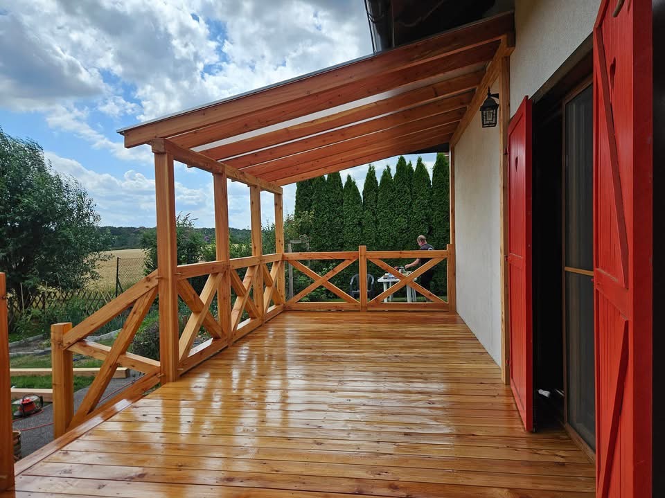 Zadaszony taras z jasnego drewna z balustradą w kształcie krzyży, świeżo polakierowana podłoga odbija światło, w tle widoczny mężczyzna przy stoliku oraz rząd wysokich tuj.