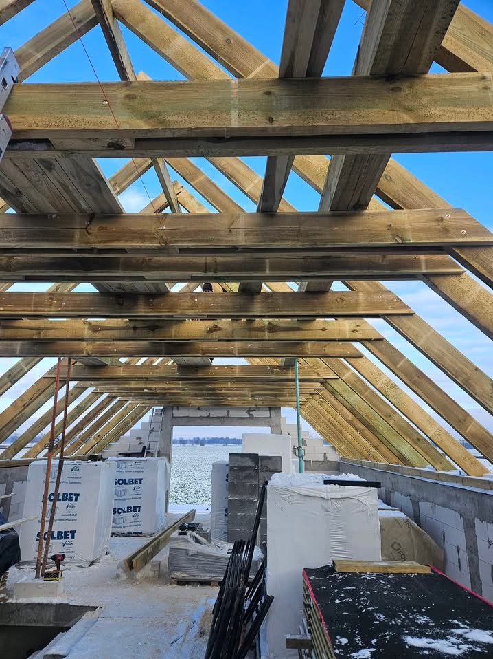 Wnętrze budynku w stanie surowym otwartym z widoczną drewnianą konstrukcją dachu, bloczkami SOLBET i oknem z widokiem na zaśnieżone pole.