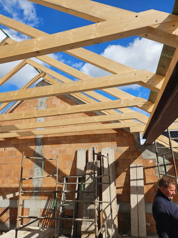 Wznoszenie drewnianej konstrukcji dachu z widoczną więźbą i elementami murowanej ściany z pustaków ceramicznych, na tle błękitnego nieba z chmurami, z widocznymi rusztowaniami i fragmentem sylwetki...