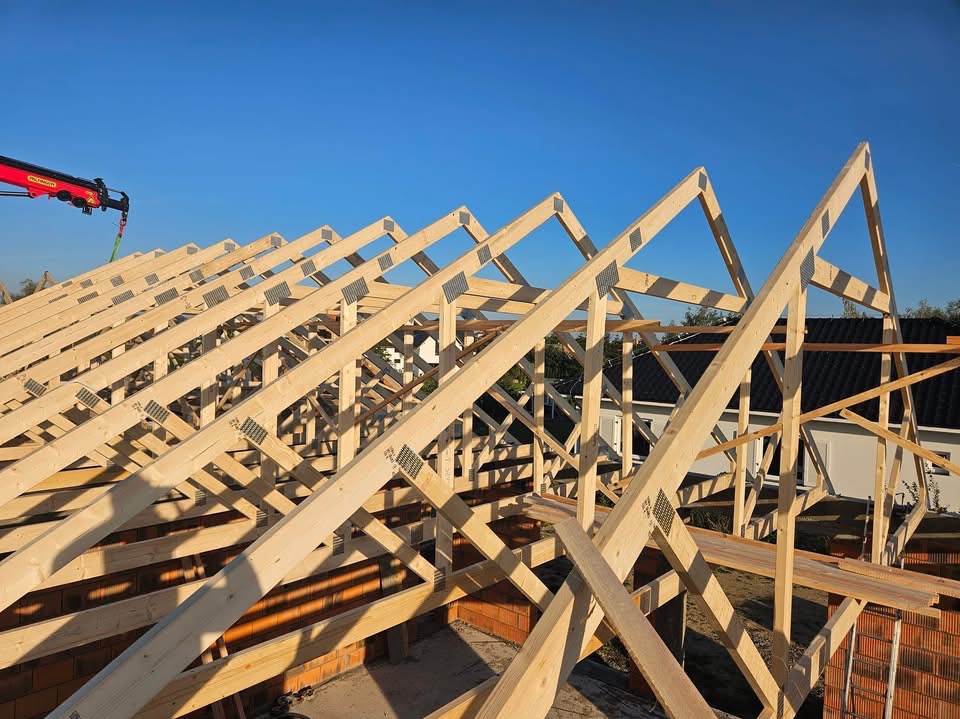 Konstrukcja drewniana dachu w trakcie montażu, widoczne elementy złączone metalowymi płytkami, na tle błękitnego nieba i częściowo wykończonego budynku.