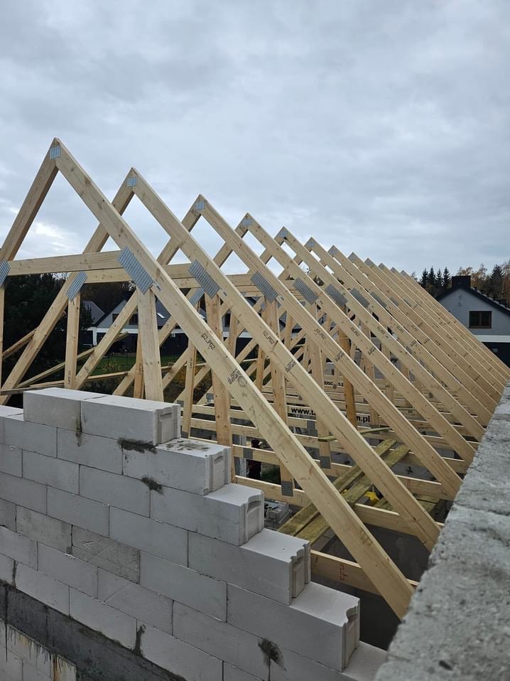 Wznoszenie drewnianej konstrukcji dachowej z prefabrykowanych elementów, widoczne metalowe łączniki na tle muru z bloczków betonowych i pochmurnego nieba.