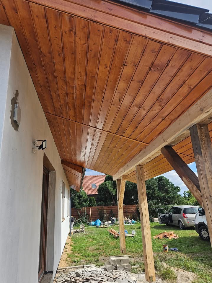 Drewniana podbitka dachowa z widoczną strukturą drewna, zamontowana na zewnątrz domu z widocznymi elementami konstrukcyjnymi tarasu oraz fragmentem elewacji i otoczenia.