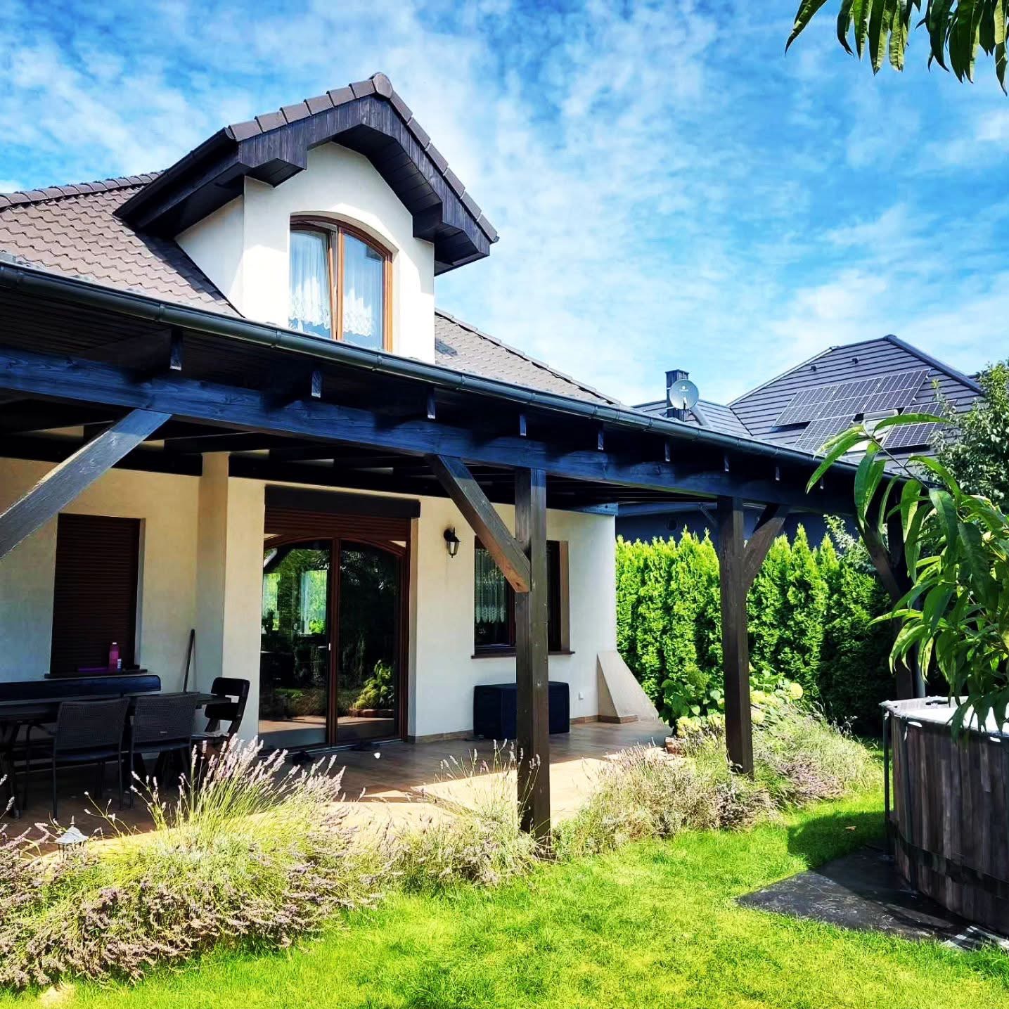 Nowoczesny dom z beżową elewacją, brązowym dachem i drewnianą pergolą, widok z ogrodu w słoneczny dzień, na dachu widoczne panele słoneczne.