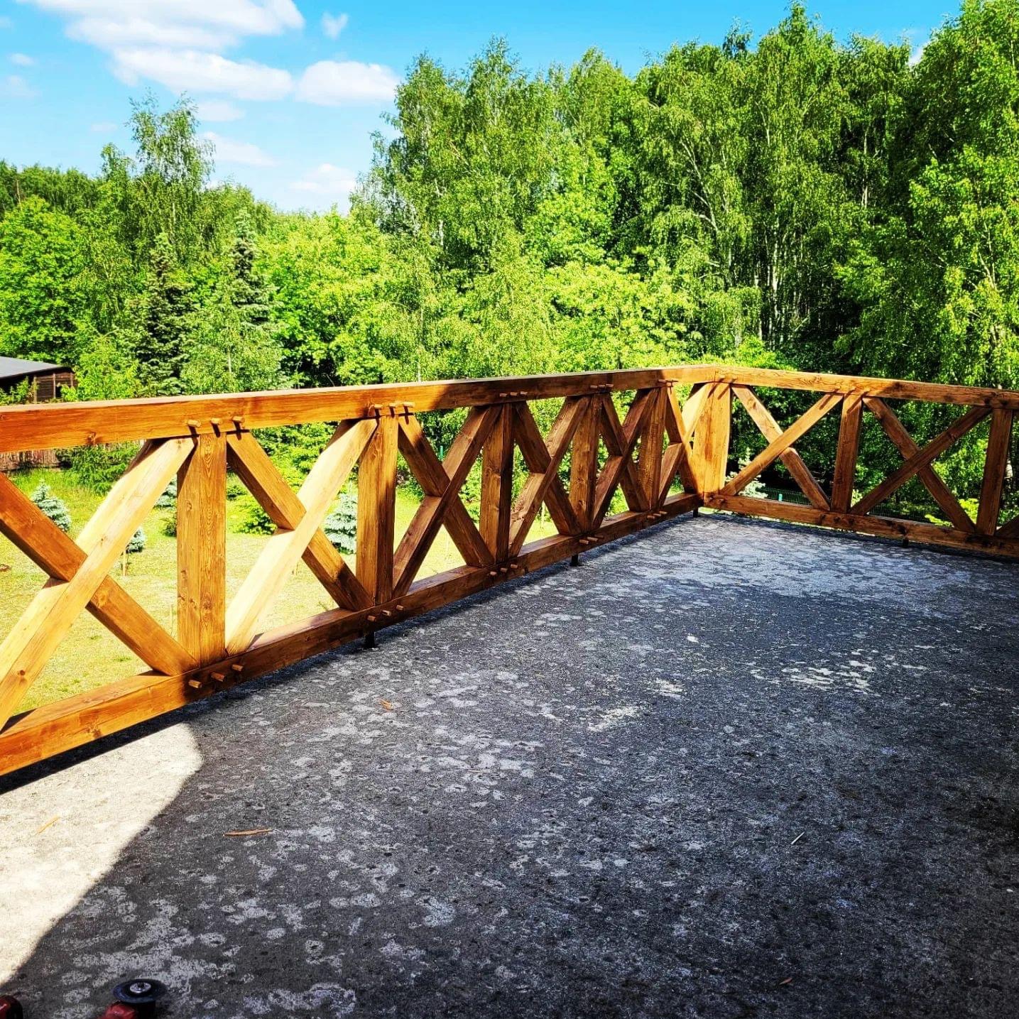 Drewniana balustrada tarasu z widokiem na bujną zieleń drzew, betonowa posadzka tarasu z widocznymi śladami użytkowania, fragment narzędzia w rogu kadru.