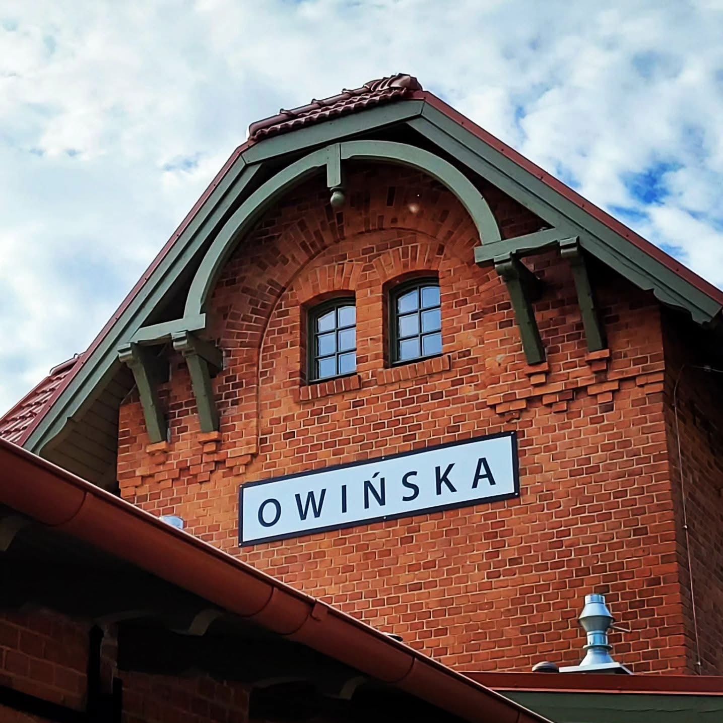 Murowana fasada budynku stacji kolejowej z dwuspadowym dachem pokrytym czerwoną dachówką, zielonymi elementami dekoracyjnymi i tablicą z nazwą stacji 'OWIŃSKA'.