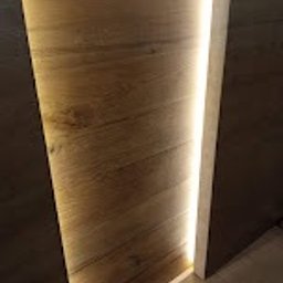 Sunny-home - Wąskie ujęcie na fragment ściany z drewnianą okładziną i pionowym paskiem oświetlenia LED wzdłuż krawędzi, widoczny fragment podłogi, ciemne tło.