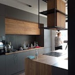 Sunny-home - Nowoczesna kuchnia z szarymi szafkami dolnymi i drewnianymi szafkami górnymi. Na blacie widoczny ekspres do kawy, naczynia i akcesoria kuchenne. Nad wyspą kuchenną metalowa konstrukcja...