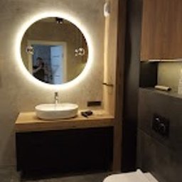 Sunny-home - Nowoczesna łazienka z okrągłym lustrem podświetlanym LED, białą umywalką nablatową na drewnianym blacie, czarną szafką pod umywalką i wiszącą toaletą w kolorze białym. Ściana wykończona w szarym...