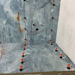 Sunny-home - Niebiesko-szara łazienka z marmurowym wzorem w trakcie wykańczania, z użyciem pomarańczowych klipsów poziomujących płytki na ścianach i podłodze, widoczne otwory na armaturę.