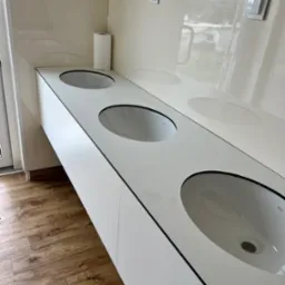 Biała, długa umywalka z trzema owalnymi zlewami marki Rea, zamontowana w łazience z panelami podłogowymi i kremowymi ścianami z dwoma włącznikami światła.