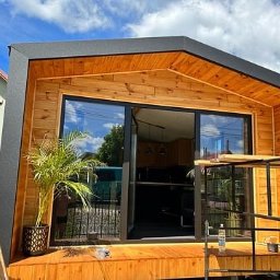 Sunny-home - Nowoczesna, drewniana altana z przesuwnymi drzwiami szklanymi, widok z zewnątrz na częściowo wykończony taras z rośliną doniczkową i butelką wody.