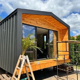 Sunny-home - Nowoczesny, mobilny dom z czarną elewacją i drewnianym wykończeniem, częściowo na paletach, z palmą w doniczce przy wejściu i rozstawioną drabiną, na tle błękitnego nieba i zieleni.