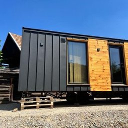 Sunny-home - Mobilny, nowoczesny dom modułowy z drewnianą elewacją i czarnymi panelami, osadzony na paletach, na tle wiejskiego krajobrazu z drewnianym budynkiem i błękitnym niebem.