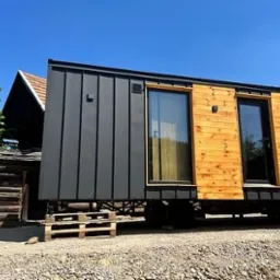 Mobilny, nowoczesny dom modułowy z drewnianą elewacją i czarnymi panelami, osadzony na paletach, na tle wiejskiego krajobrazu z drewnianym budynkiem i błękitnym niebem.