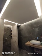 Nowoczesna łazienka z szarymi ścianami imitującymi beton, podświetlany sufit z ukrytymi listwami LED, biała umywalka nablatowa na drewnianym blacie, kabina prysznicowa typu walk-in z szarą armaturą...