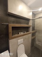 Narożna półka z surowego drewna w nowoczesnej łazience z szarymi płytkami, widoczna poziomica wewnątrz półki, toaleta w dolnej części kadru.