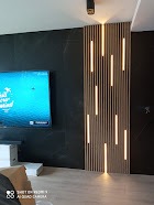 Nowoczesny salon z ciemną ścianą, panelem dekoracyjnym z pionowych listew z podświetleniem LED i telewizorem na ścianie.