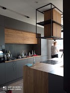 Nowoczesna kuchnia z szarymi szafkami dolnymi i drewnianymi szafkami górnymi. Na blacie widoczny ekspres do kawy, naczynia i akcesoria kuchenne. Nad wyspą kuchenną metalowa konstrukcja...