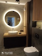 Nowoczesna łazienka z okrągłym lustrem podświetlanym LED, białą umywalką nablatową na drewnianym blacie, czarną szafką pod umywalką i wiszącą toaletą w kolorze białym. Ściana wykończona w szarym...