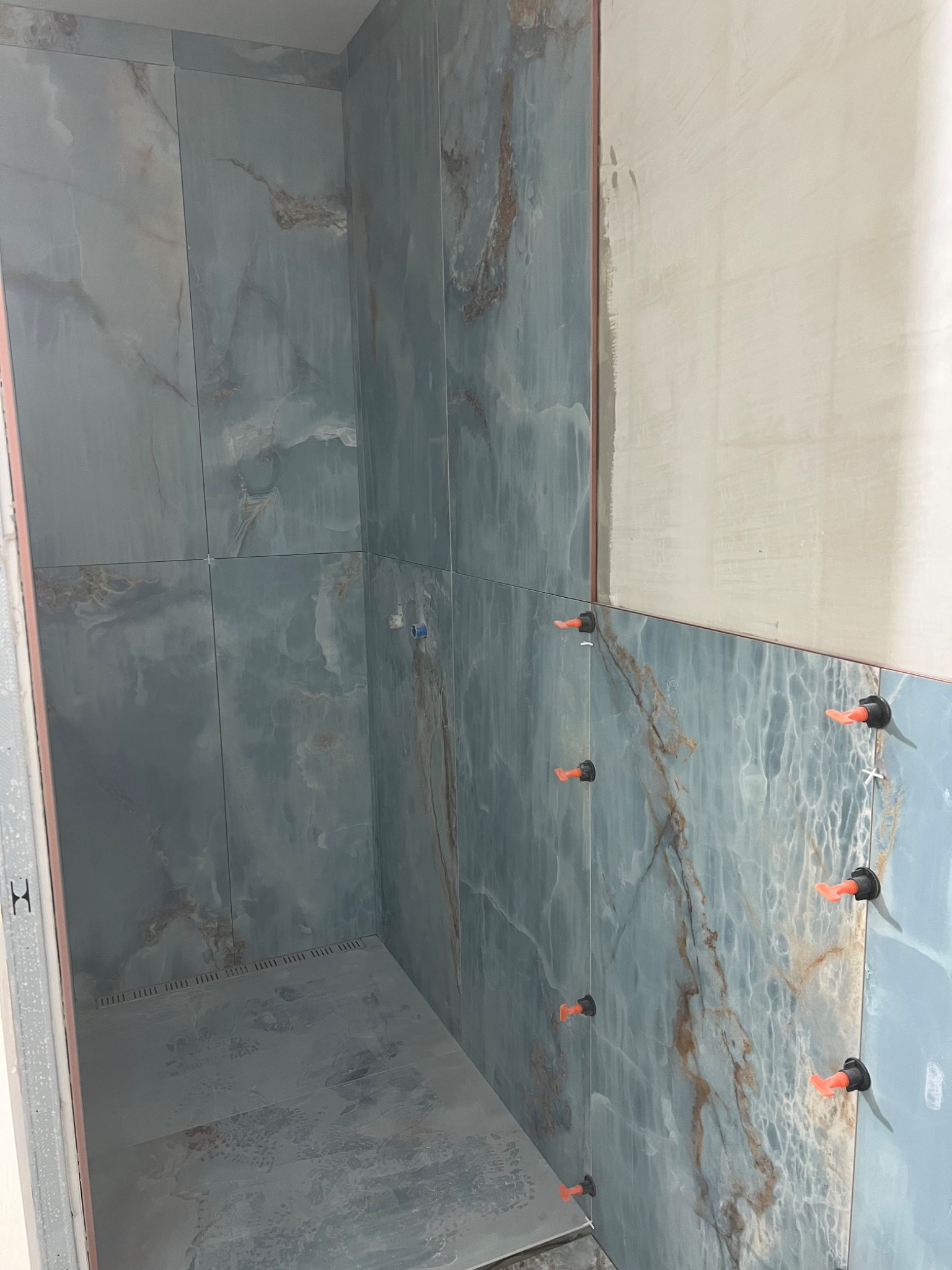 Montaż płytek imitujących marmur w kabinie prysznicowej, z użyciem pomarańczowych klipsów poziomujących, widoczna surowa ściana przygotowana do dalszych prac.