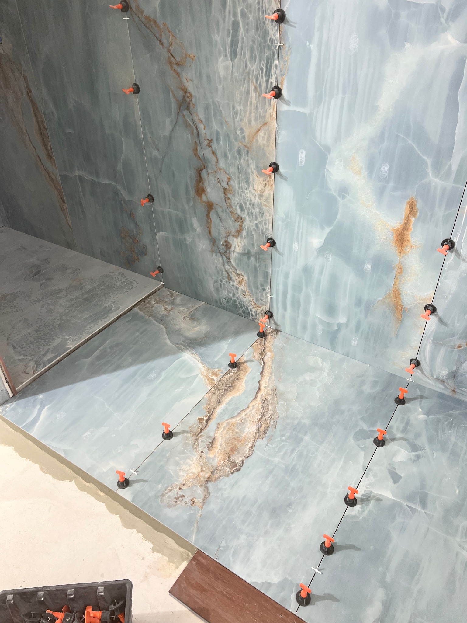 Ściany i podłoga w trakcie wykańczania płytkami imitującymi onyks, z użyciem systemu poziomowania płytek z pomarańczowymi klipsami i czarnymi podstawkami.