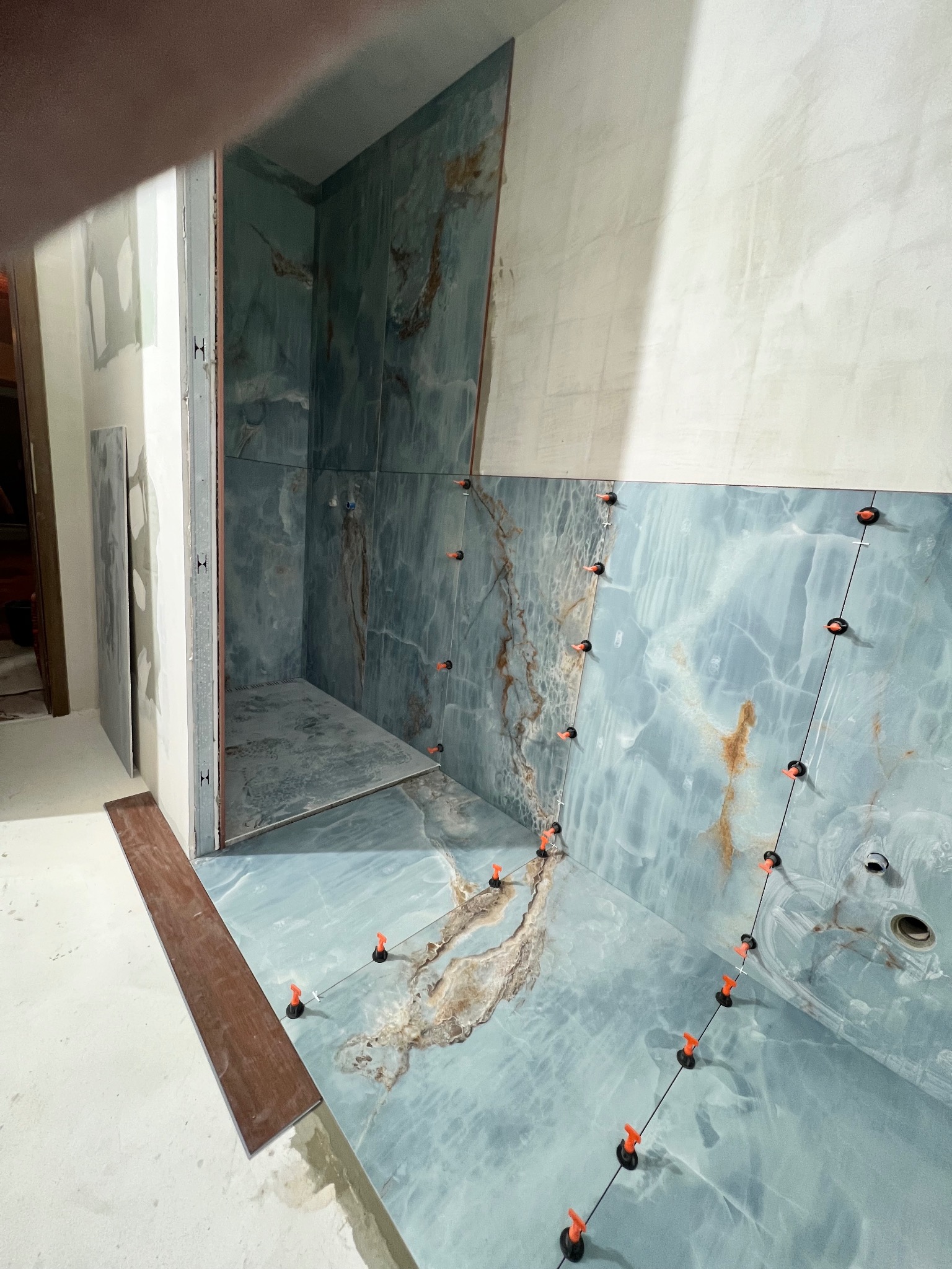 Niebiesko-marmurowe płytki ścienne w trakcie montażu w łazience, widoczne pomarańczowe klipsy poziomujące i czarne przyssawki, surowa ściana w tle.