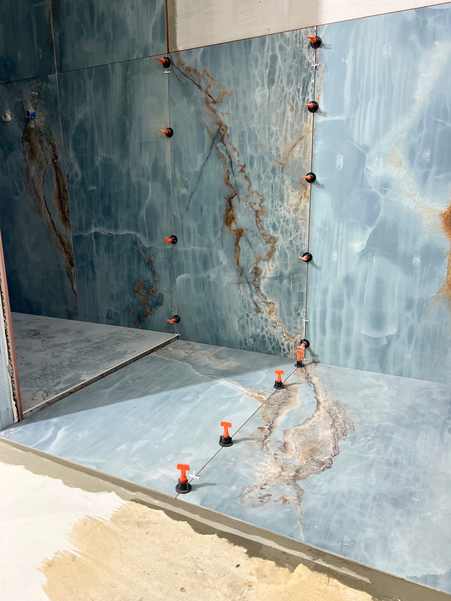 Ściana i podłoga w trakcie wykańczania, wyłożone błękitnymi płytkami imitującymi marmur, z pomarańczowymi elementami poziomującymi na powierzchni.