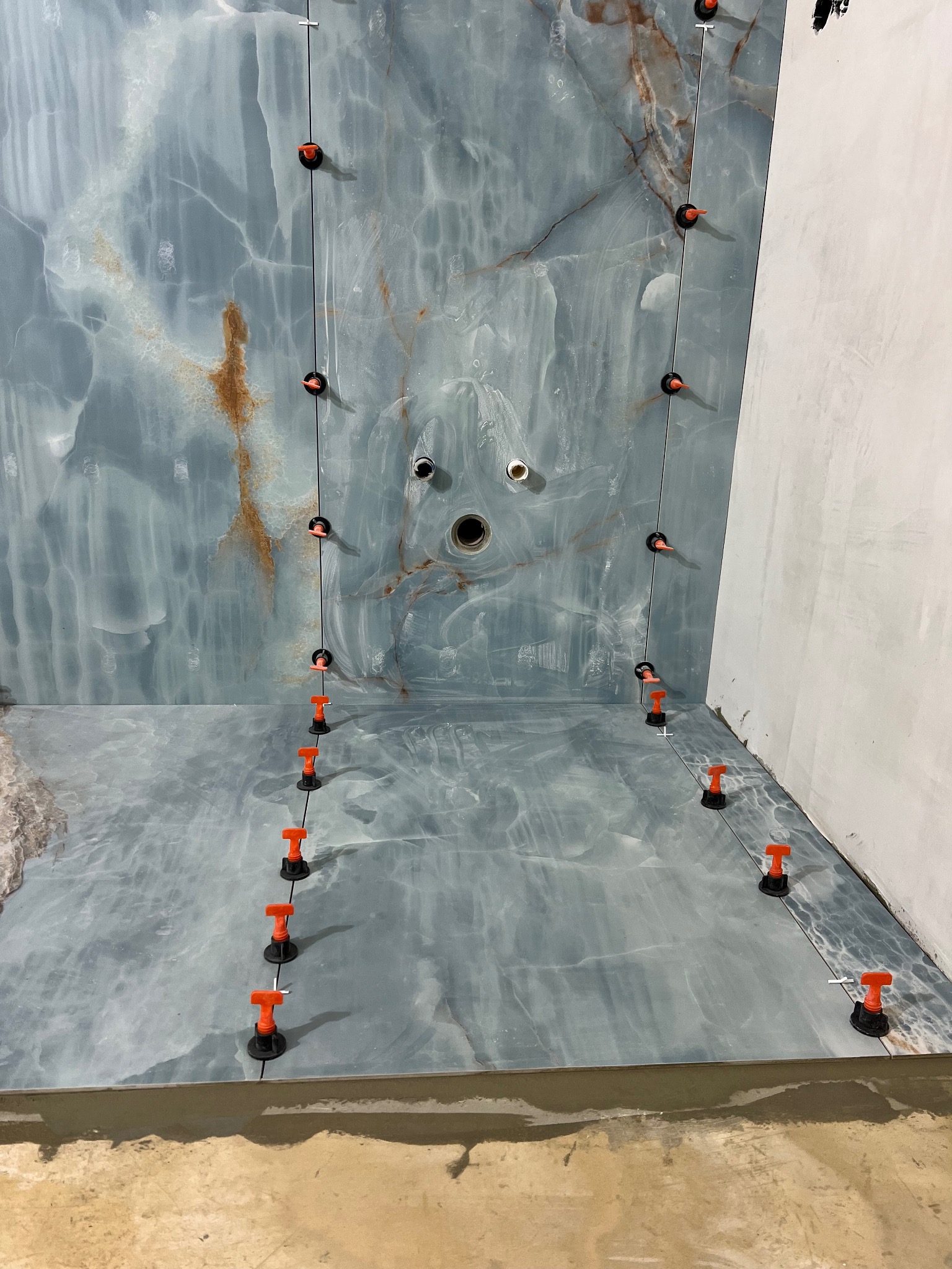 Niebiesko-szara łazienka z marmurowym wzorem w trakcie wykańczania, z użyciem pomarańczowych klipsów poziomujących płytki na ścianach i podłodze, widoczne otwory na armaturę.