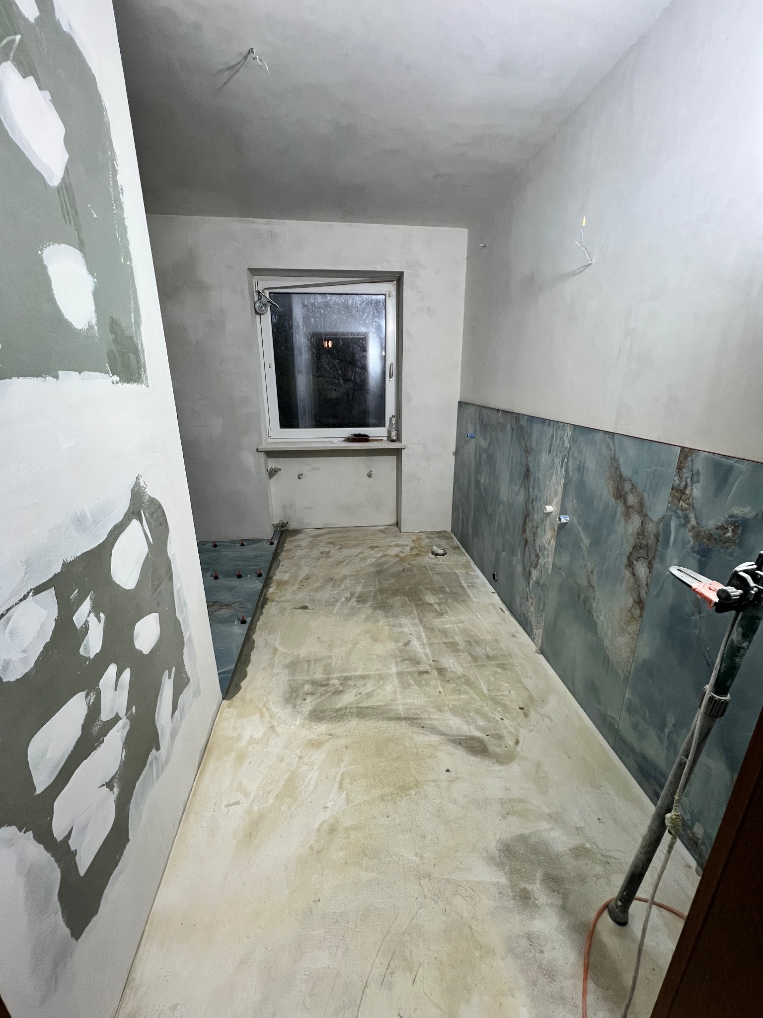 Łazienka w trakcie remontu: ściany częściowo wyłożone marmurowymi płytkami w odcieniach szarości i błękitu, przygotowane podłoże pod płytki na podłodze, widoczna poziomica, ściana z nałożoną...