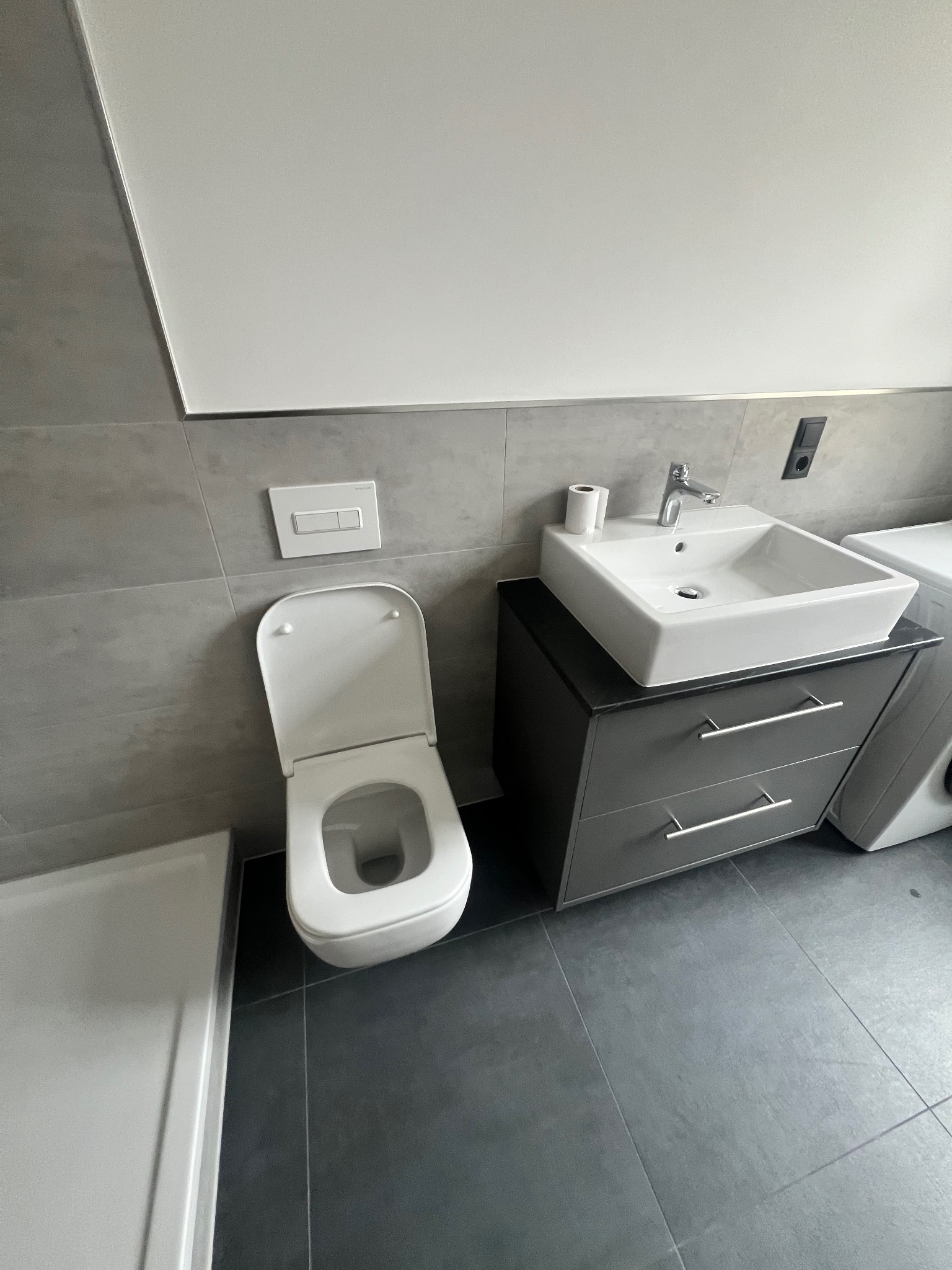 Nowoczesna łazienka z szarą glazurą, wiszącą toaletą z otwartą klapą, umywalką nablatową na szafce z szufladami i fragmentem brodzika.