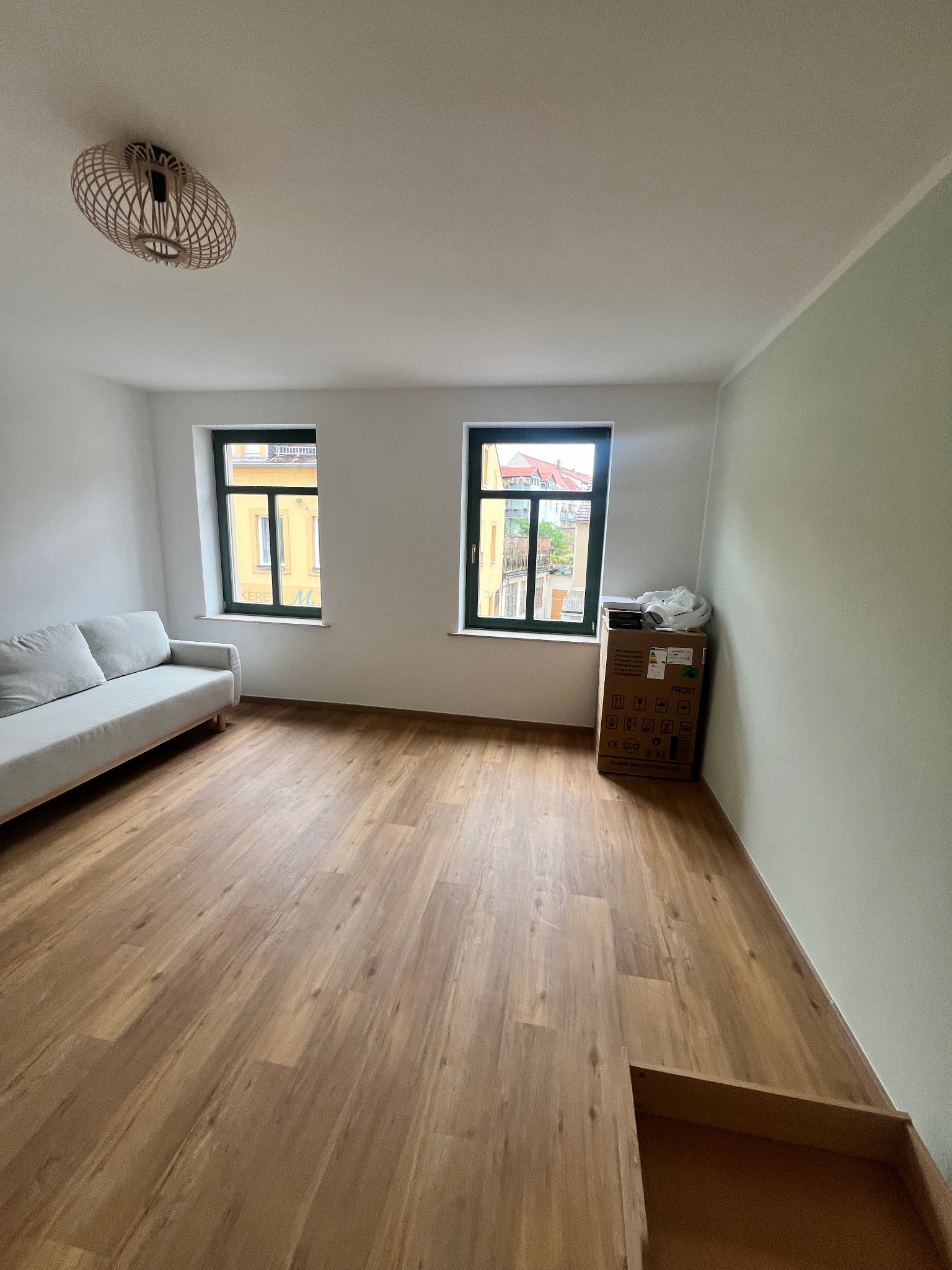 Jasne wnętrze pokoju po remoncie z nową podłogą z paneli, świeżo pomalowaną ścianą w odcieniach zieleni, szarą sofą i oknami z zielonymi ramami. Na podłodze stoi karton z akcesoriami.