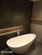 Nowoczesna, wolnostojąca wanna owalna z chromowaną baterią w minimalistycznej łazience w Tarnowie. Ciemne, strukturalne płytki na ścianie kontrastują z jasnym drewnem półki i bielą wanny.