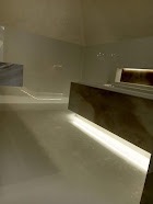 Minimalistyczna łazienka z podświetlanymi elementami, jasnoszara posadzka żywiczna, brak tradycyjnych płytek, surowe wykończenie, oświetlenie punktowe.