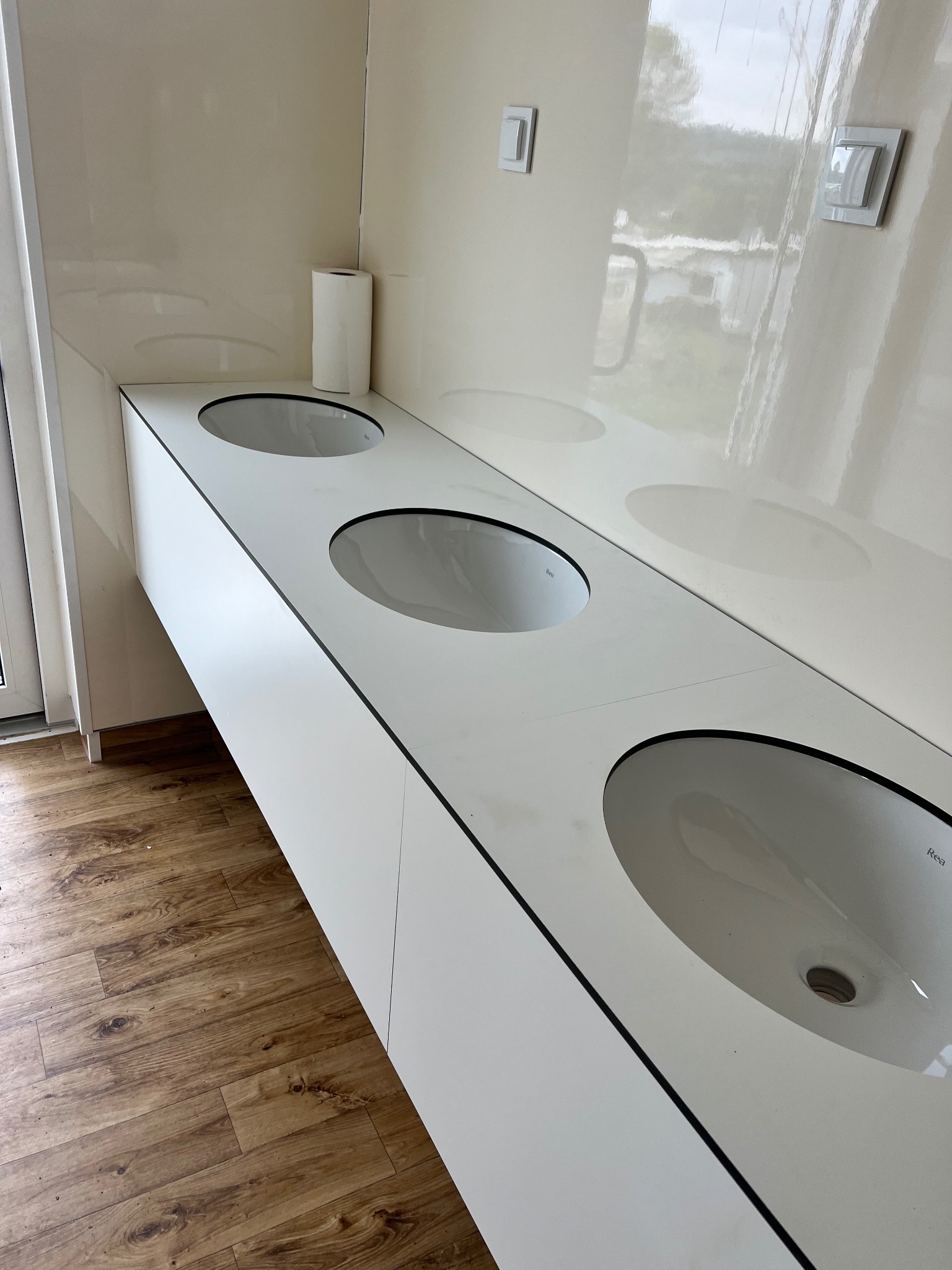 Biała, długa umywalka z trzema owalnymi zlewami marki Rea, zamontowana w łazience z panelami podłogowymi i kremowymi ścianami z dwoma włącznikami światła.