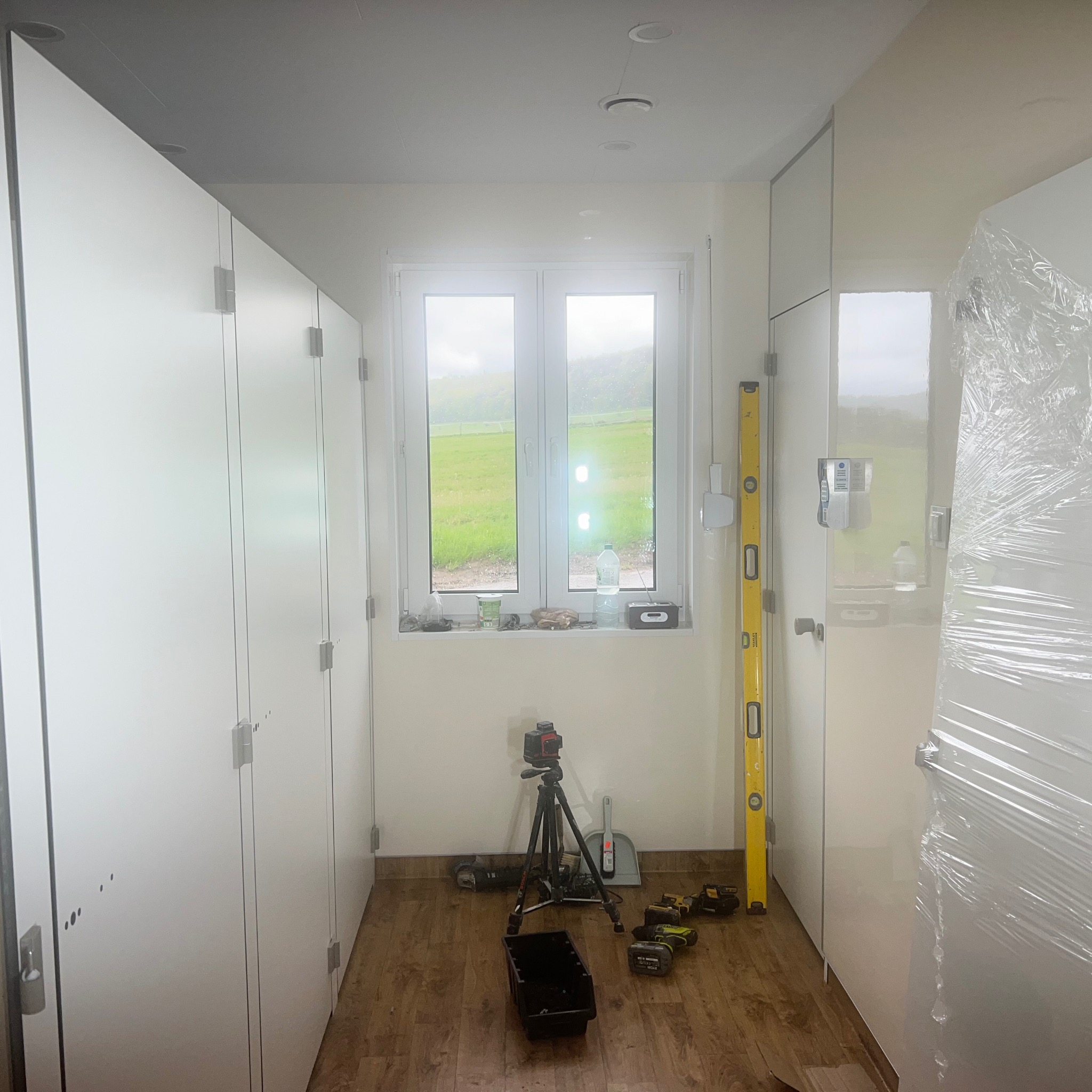 Wnętrze pomieszczenia z białymi szafami w zabudowie, okno z widokiem na zielony krajobraz, poziomica oparta o szafę, laser krzyżowy na statywie i narzędzia na podłodze z paneli.