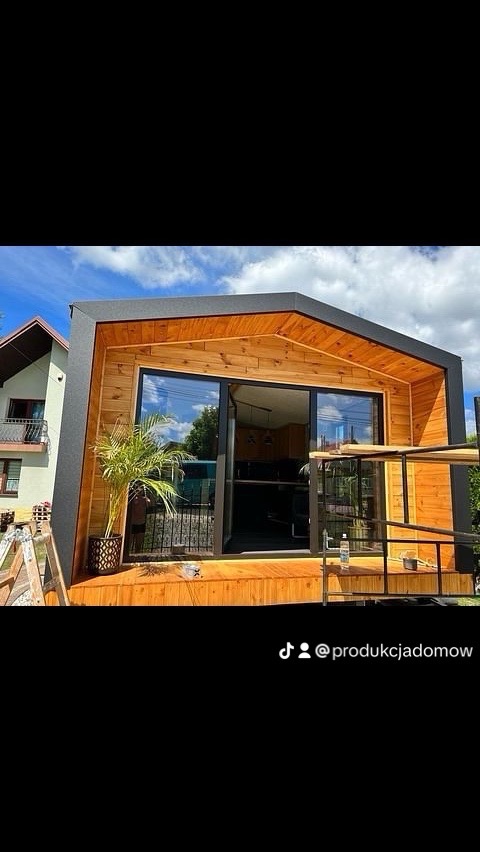 Nowoczesna drewniana konstrukcja altany z przesuwnymi drzwiami, widok z zewnątrz, z rośliną doniczkową i fragmentem drabiny budowlanej po lewej stronie.